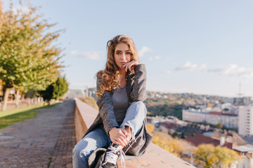 Serious well-dressed girl with curly hair propping face with hand. Lovely young woman in blue jeans...