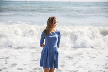 Fototapeta na wymiar Beautiful girl in blue dress is walking on the beach. Amazing summer photo. Woman near the sea. Happy and fun emotions. Holiday travel concept. Slim legs. Warm ocean water.