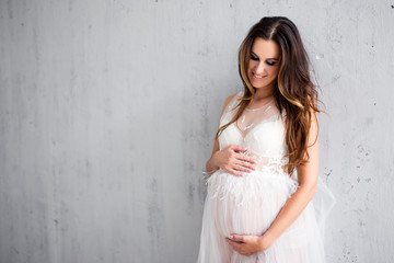 Fototapeta na wymiar beautiful pregnant woman in white lace dress posing over gray wall background with copy space