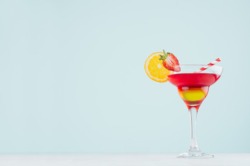 Sunrise cocktail with red and yellow liquor, orange slice, strawberry, straw on elegant white wood board and pastel mint color wall.