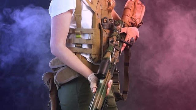 Woman soldier in military camouflage uniform protected with headphones, body armour, holding assault rifle standing against dark smoky background