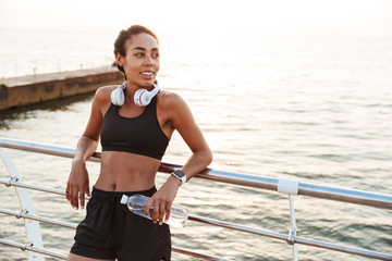 Image of smiling pretty woman bending over railings and holding water bottle while standing by...