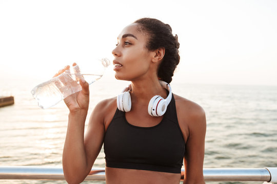 Image of beautiful woman with headphones bending over railings and drinking water by seaside in morning