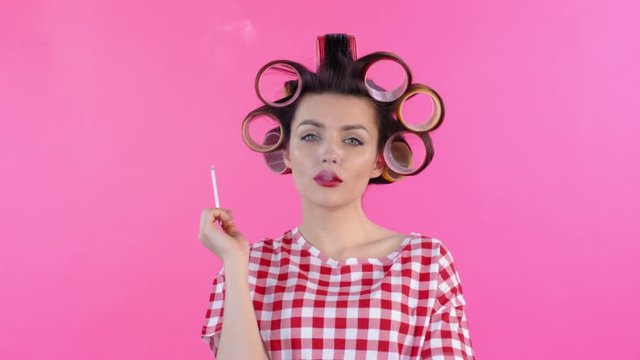 Medium shot of attractive young Caucasian woman standing on pink background and smoking cigarette while curling hair with hair rollers
