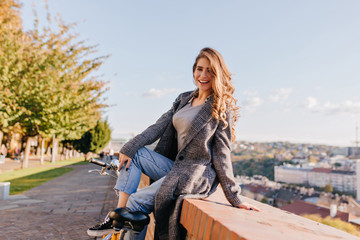 Inspired young female model in gray coat posing in park after riding on bicycle and smiling. Fashionable woman in blue jeans sitting on the street with amazing city view behind.