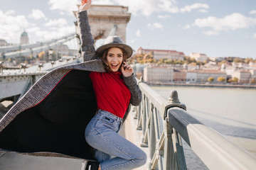 Excited long-haired woman dancing in long coat and laughing on urban background. Slim girl in red...