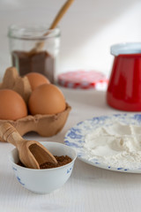 Vertical top view of ingredients for pastries, brown sugar, flour, eggs and red saucepan on white wooden background