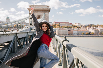 Carefree girl in blue jeans happy dancing on architecture background. Enthusiastic curly female model expressing positive emotions in weekend in Europe.