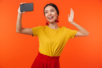 Happy young woman posing isolated over orange wall background talking by mobile phone take a selfie.