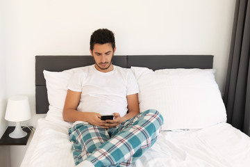 Young handsome man sitting on the bed. He uses a mobile phone. The problem of dependence on mobile social networks.