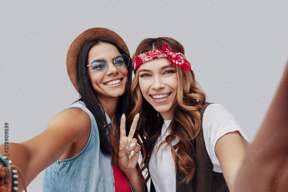 Poster Self portrait of two attractive stylish young women looking at camera and smiling while standing against grey background