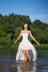 Fototapeta na wymiar Young pretty brunette woman in white wedding dress, walking on the river and making splashes