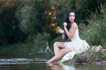 Young pretty brunette woman in white wedding dress sitting on the rock near forest river