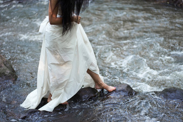 Young pretty brunette woman in white wedding dress trying to cross the river by rocks