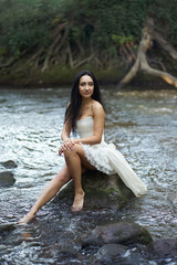 Young pretty brunette woman in white wedding dress sitting on the rock in swirling river