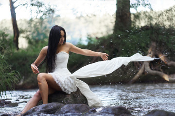 Young pretty brunette woman in white wedding dress sitting on the rock in rushing river and corrects her dress