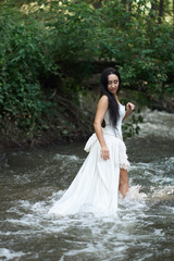 Fototapeta na wymiar Young pretty brunette woman in white wedding dress stands outdoors in rushing river