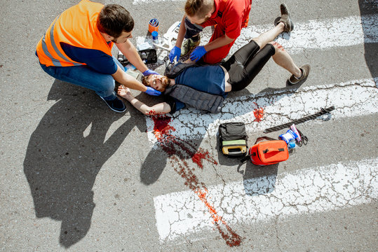 Ambulance Worker With Man In Road Vest Applying Emergency Medical Care To The Injured Bleeding Person Lying On The Pedestrian Crossing After The Accident, View From The Above