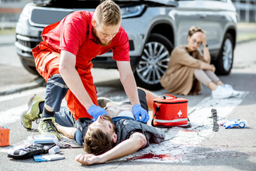 Ambluence worker applying emergency care to the injured bleeding man lying on the pedestrian crossing, dispair woman driver sitting on the background