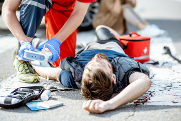 Ambluence worker applying emergency care with defibrillator to the injured bleeding man lying on the pedestrian crossing - obrazy, fototapety, plakaty