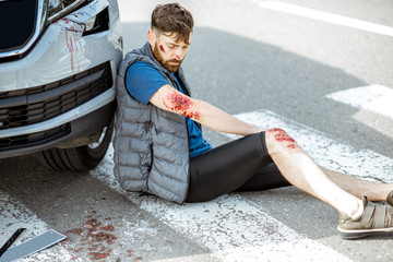 Injured man with bleeding wounds sitting on the pedestrian crossing near the car after the road...