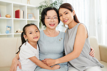 Fototapeta na wymiar Portrait of happy Asian family generation mother daughter and grandmother embracing and smiling at camera while resting on couch