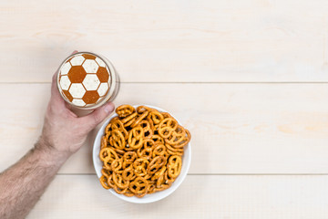 Man's hand holds a beer mug with a soccer ball on a beer foam with pretzels. Top view. Empty space for text