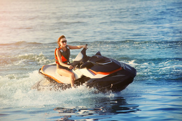 Young adult sporty caucasian woman riding jet ski in ocean blue water at warm evening sunset. Beach...