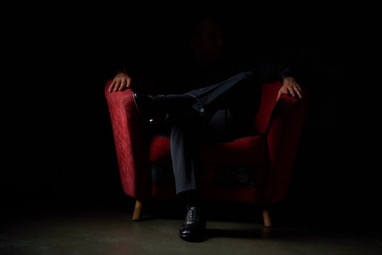 male businessman in a black suit sitting in red chair, black background, no faces visible, studio shooting