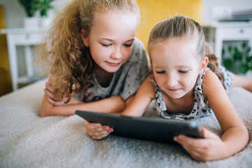 Two beautiful little sisters lying in the bed and look at the screen of a tablet, smart kids using...