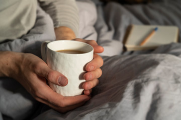 Hygge, lagom lifestyle image with man holding coffee cup. Background with open notebook and a pencil on the bed, covered with minimalist grey linen sheet. Cozy lifestyle.
