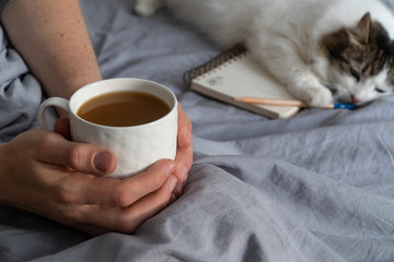 Hygge, lagom lifestyle image with man holding coffee cup. Background with cute cat, open notebook and a pencil on the bed, covered with minimalist grey linen sheet. Cozy lifestyle.