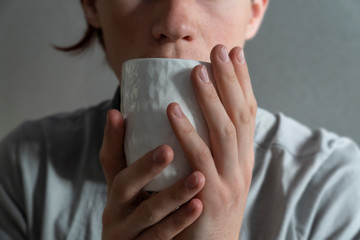 Hygge, lagom lifestyle image with a man holding a coffee cup in front of his face. Man taking a sip of a hot drink from a white cup. Minimalist grey background. Cozy lifestyle.
