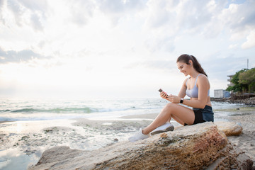 Fototapeta na wymiar Pretty brunette woman with smart watches reading messages on the mobile phone at the sea shore after morning workout at sunrise while listening to the music with wireless earbuds.