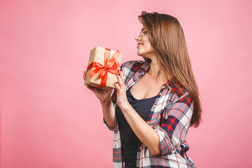 Happy young woman in casual holding gift box and looking aside while enjoys over pink background