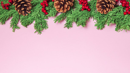 Green foam Christmas tree branch border with cones and red berries on the pink background flat lay, wide format. Handmade New Year decor with copy space, mock up