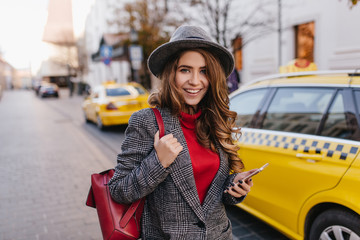 Glamorous business lady in red sweater posing with charming smile on avenue. Outdoor photo of...