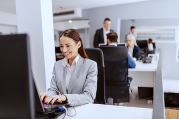 Beautiful positive Caucasian businesswoman in formal wear sitting in office and using computer. Hands on keyboard. In background colleagues working.