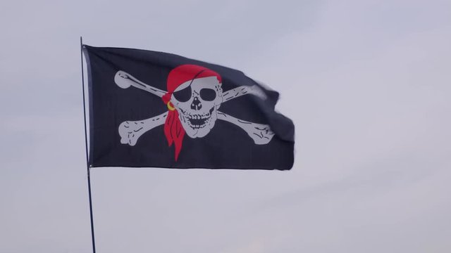 Pirate flag Jolly Roger hanging on a ship mast on a blue sky background at windy day.