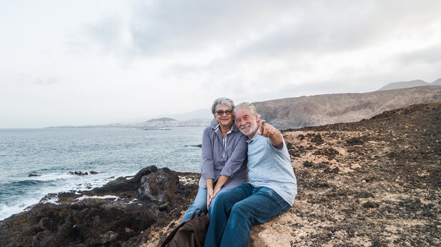 Happy senior couple of people waiting the sunrise sitting on the cliff looking at camera. Early morning for two retired person.