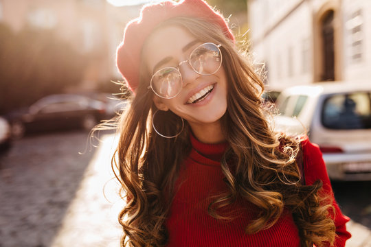 Close-up portrait of lovable french girl with elegant curly hairstyle looking to camera and laughing. Enthusiastic lovely lady in beret enjoying sunshine, walking on avenue.