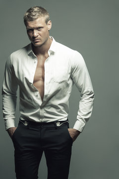 Male fashion, beauty concept. Portrait of brutal young man with short wet blond hair wearing white shirt, black pants, posing over gray background. Classic style. Studio shot