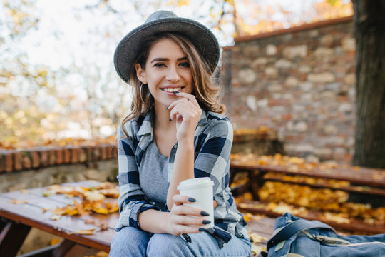 Ecstatic white woman in casual shirt holding cup of tea on blur autumn background. Outdoor photo of interested european lady in hat with long black nails smiling in october weekend.