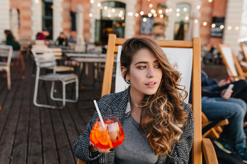 Magnificent pale woman with light-brown hair looking away with serious face expression and drinking cocktails. Portrait of bored young lady in tweed jacket spending time in outdoor cafe.
