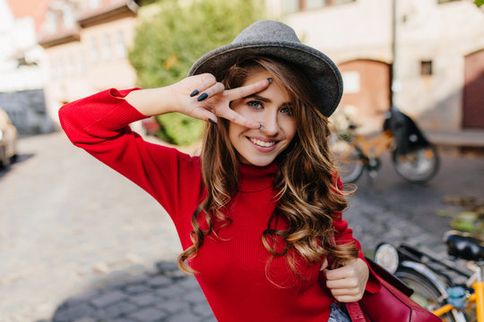 Close-up portrait of inviting blue-eyed woman with black manicure happy smiling on street background. Amazing white girl wears trendy hat posing with peace sign.