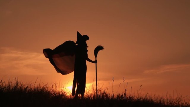 Silhouette of a witch in a raincoat and a hat at sunset she performs a ritual dance. In the hands of holding a flying broom.