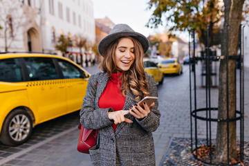 Good-looking business woman texting message while walking on the street. Refined lady in elegant coat looking at phone screen, standing on avenue with taxi on background.