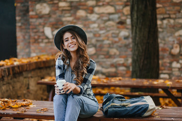 Lovable european female model wears casual jeans and blue shirt sitting outdoor in warm october weekend. Stunning young woman relaxing in park in autumn day and cute smiling to camera.