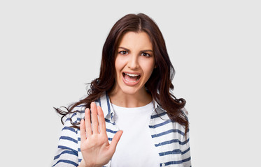 Pretty young woman making stop gesture with her hand. Portrait of Caucasian female wearing striped shirt and white t-shirt, making rejection gesture, posing on white background. Negative emotion