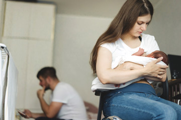A mother with a newborn baby in her arms is breastfeeding him. Father sits alone in the background...
